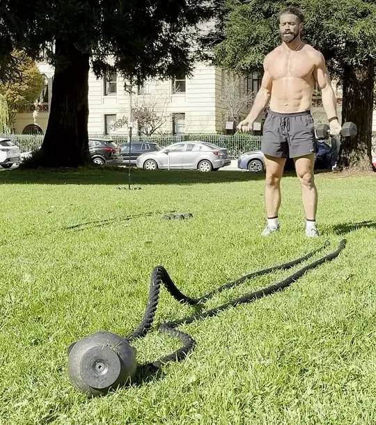 Dom curling dumbbells on the grass in a park, a battle rope laid out in front of him